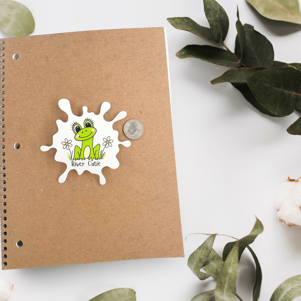 River Cutie frog vinyl sticker on a brown spiral notebook with a coin for scale.
