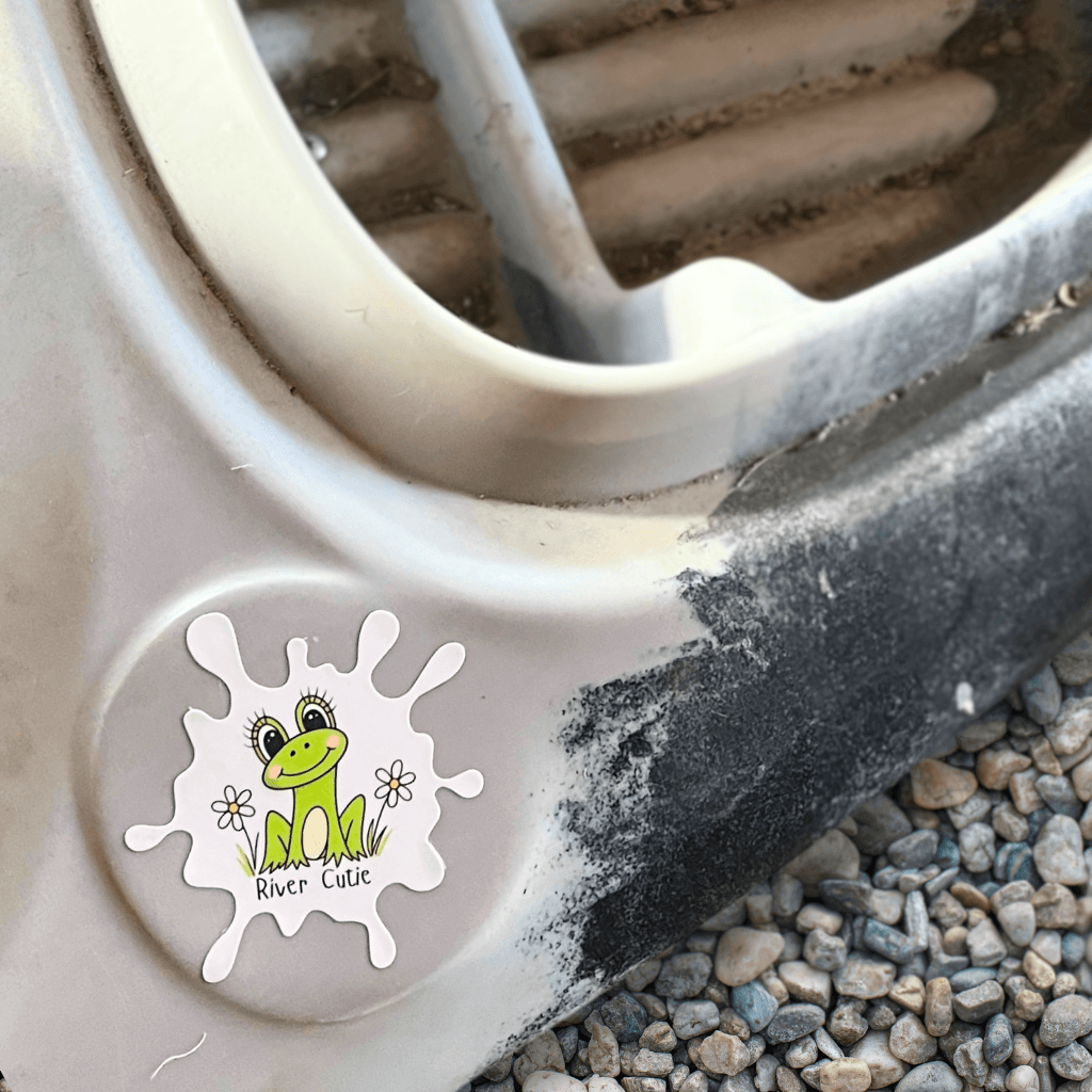 River Cutie frog sticker on a white kayak surrounded by gravel.