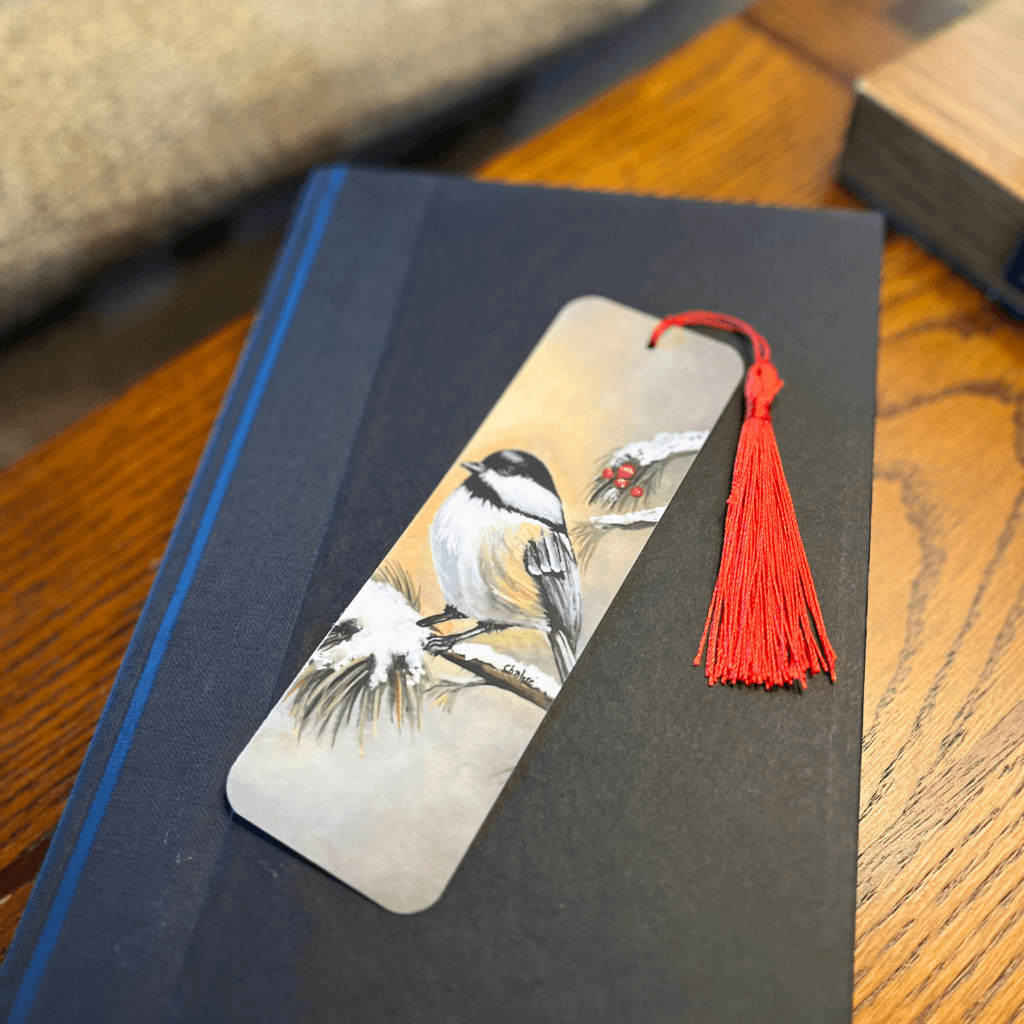 Handmade Winter Chickadee bookmark with red tassel, placed on black book cover.