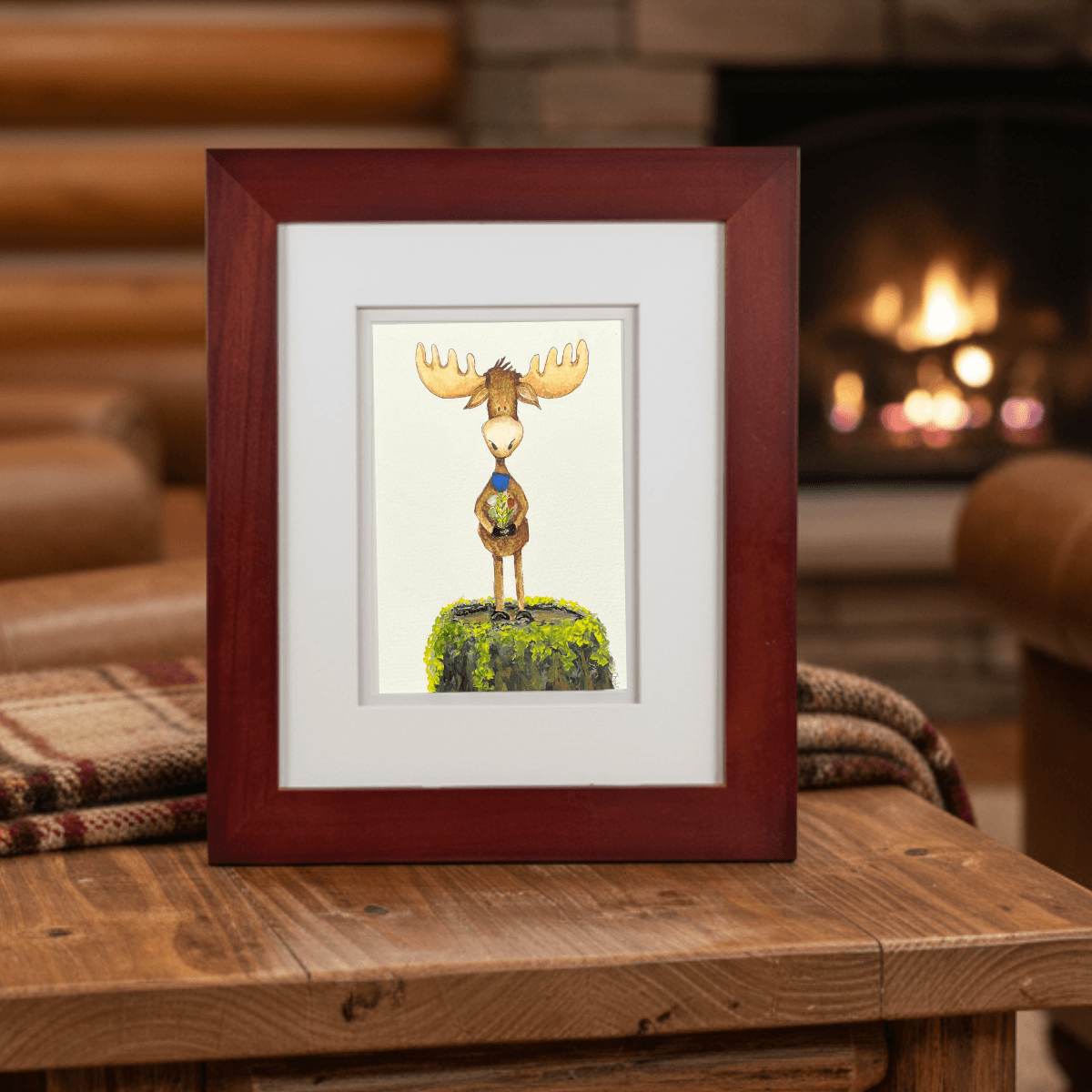 Framed You’re My Maine Squeeze moose illustration displayed on a wooden table in a cozy log cabin with a glowing fireplace in the background.