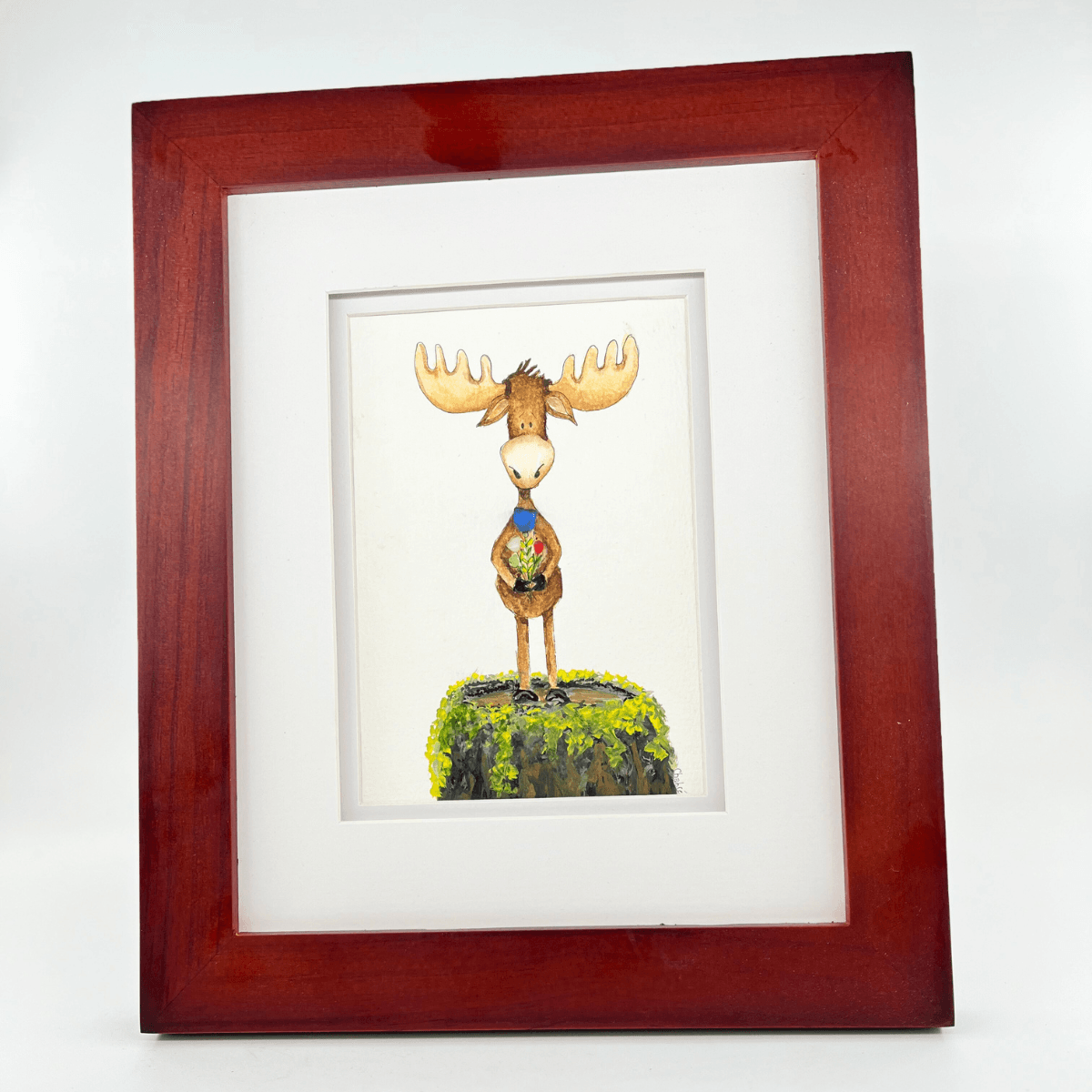 Angled view of You’re My Maine Squeeze, an original mixed media moose illustration by Riverbed Art featuring Aroostook River glass details, framed in wood with a white mat.