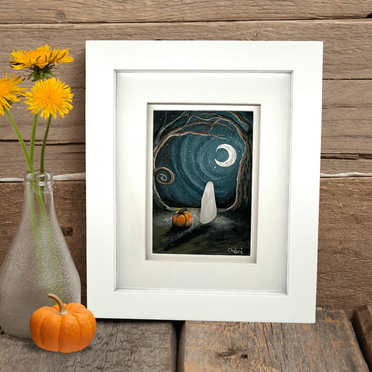 Framed gouache artwork October Night by Riverbed Art, featuring a ghost made from handpicked Aroostook River beach glass beside a pumpkin under a moonlit sky, displayed on rustic wood with flowers and a mini pumpkin.