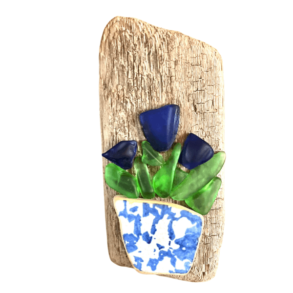 Small driftwood wall art featuring blue river glass flowers and green leaves arranged in a blue-and-white pottery shard planter