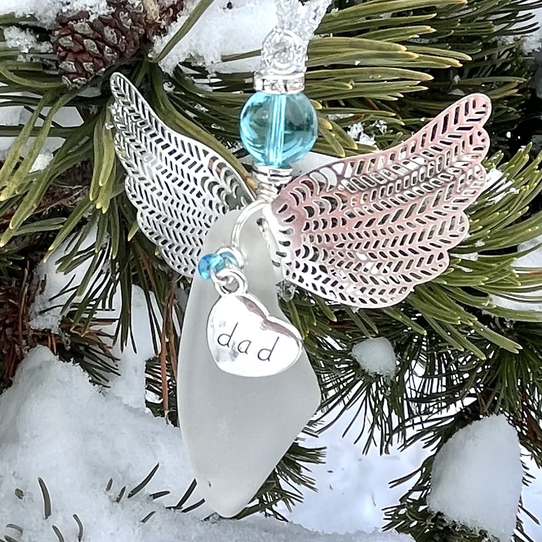 Beach glass angel ornament with silver wings and “dad” charm displayed outdoors on snowy pine branches.