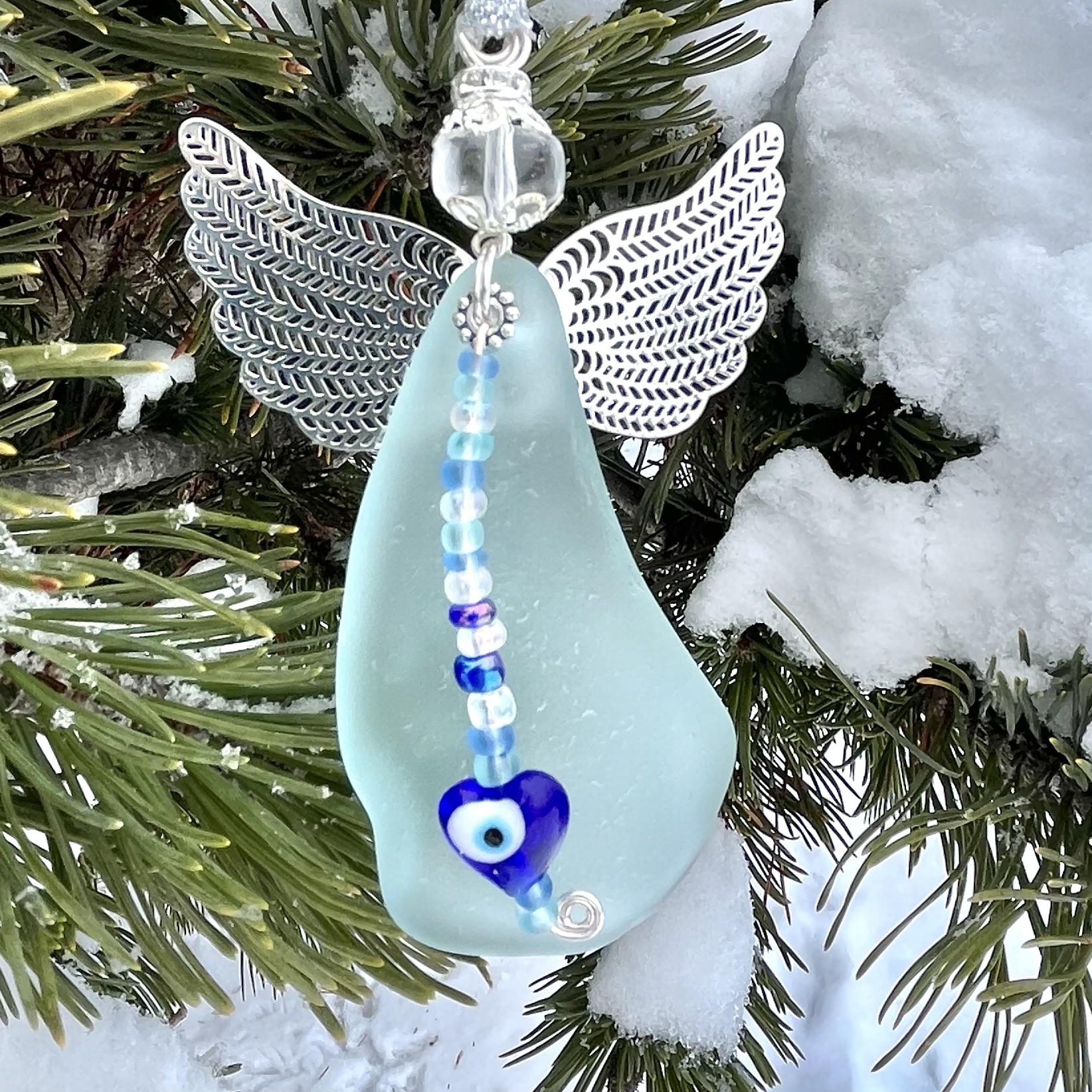 Icy-blue beach glass angel with silver-colored wings, clear glass bead head, and blue glass heart charm hanging on snowy pine branches.