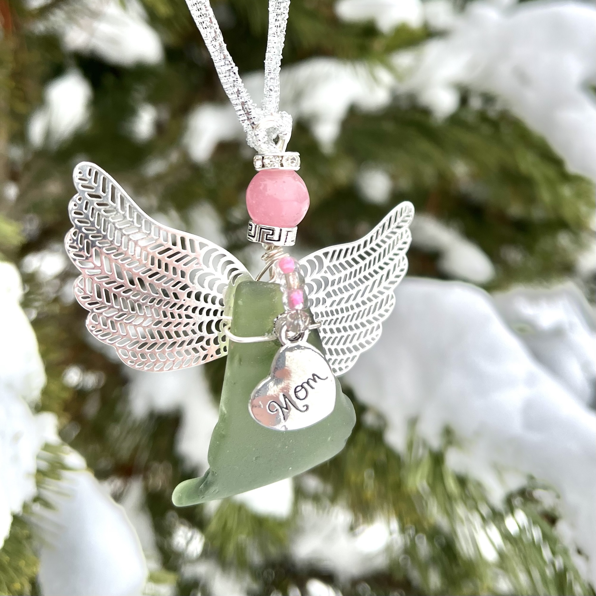 Green beach glass angel with silver wings, pink bead, and “Mom” heart charm hanging outdoors in snowy pine branches.