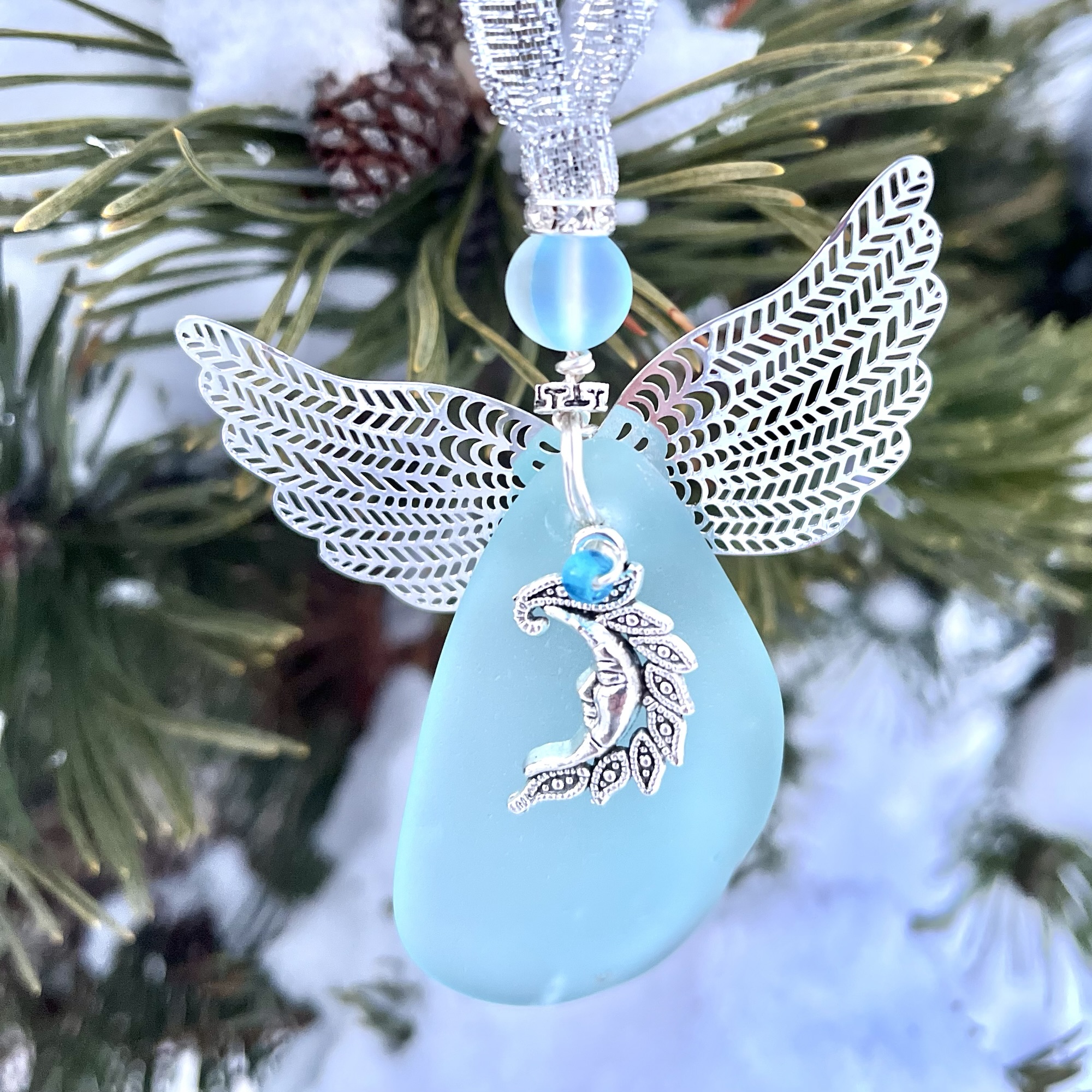Aqua beach glass angel ornament with silver wings, frosted bead, and a moon goddess charm hanging on snowy pine branches.