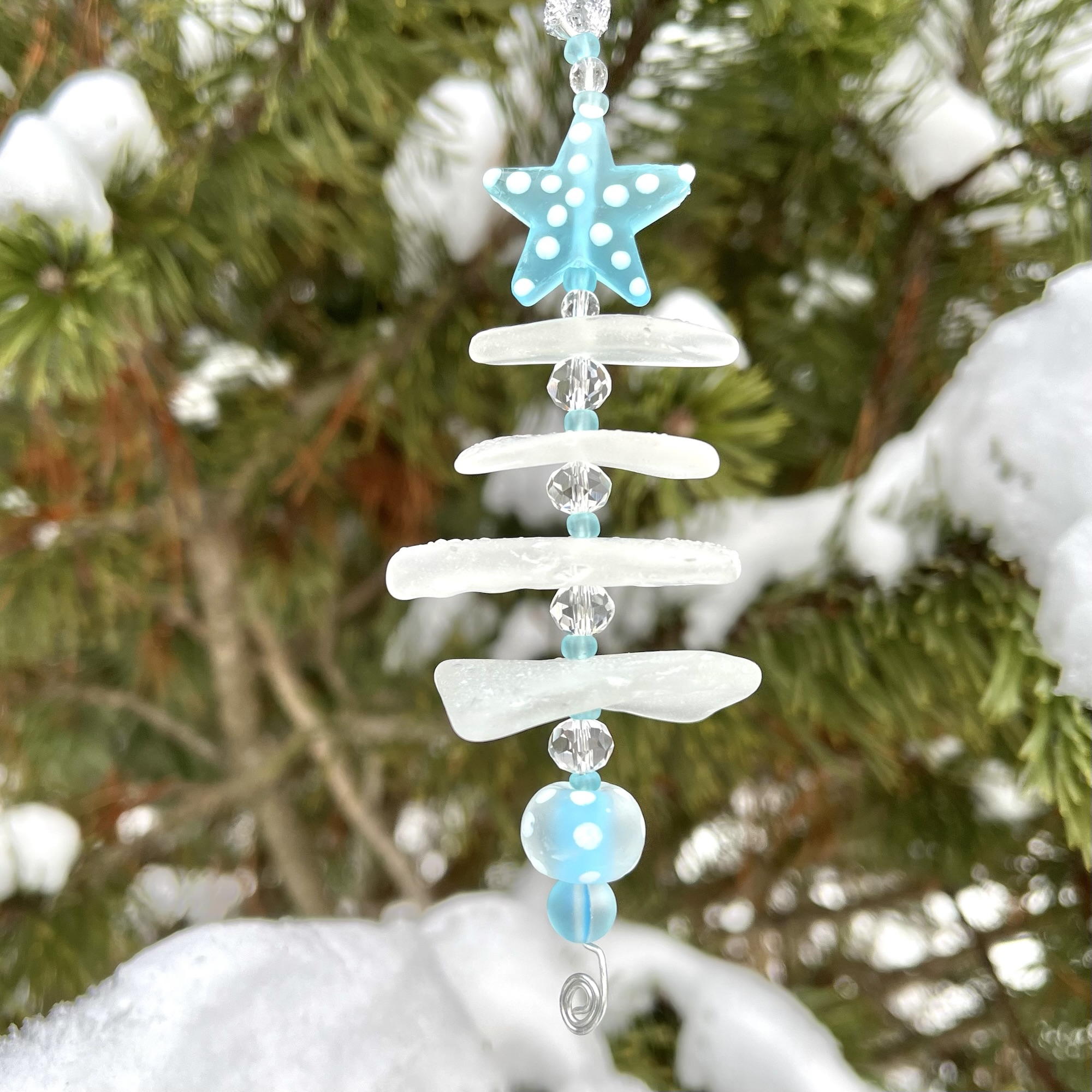 2025 Aroostook River Snow Tree ornament made from stacked frosted river glass with a blue star topper hanging outdoors in snowy pines.
