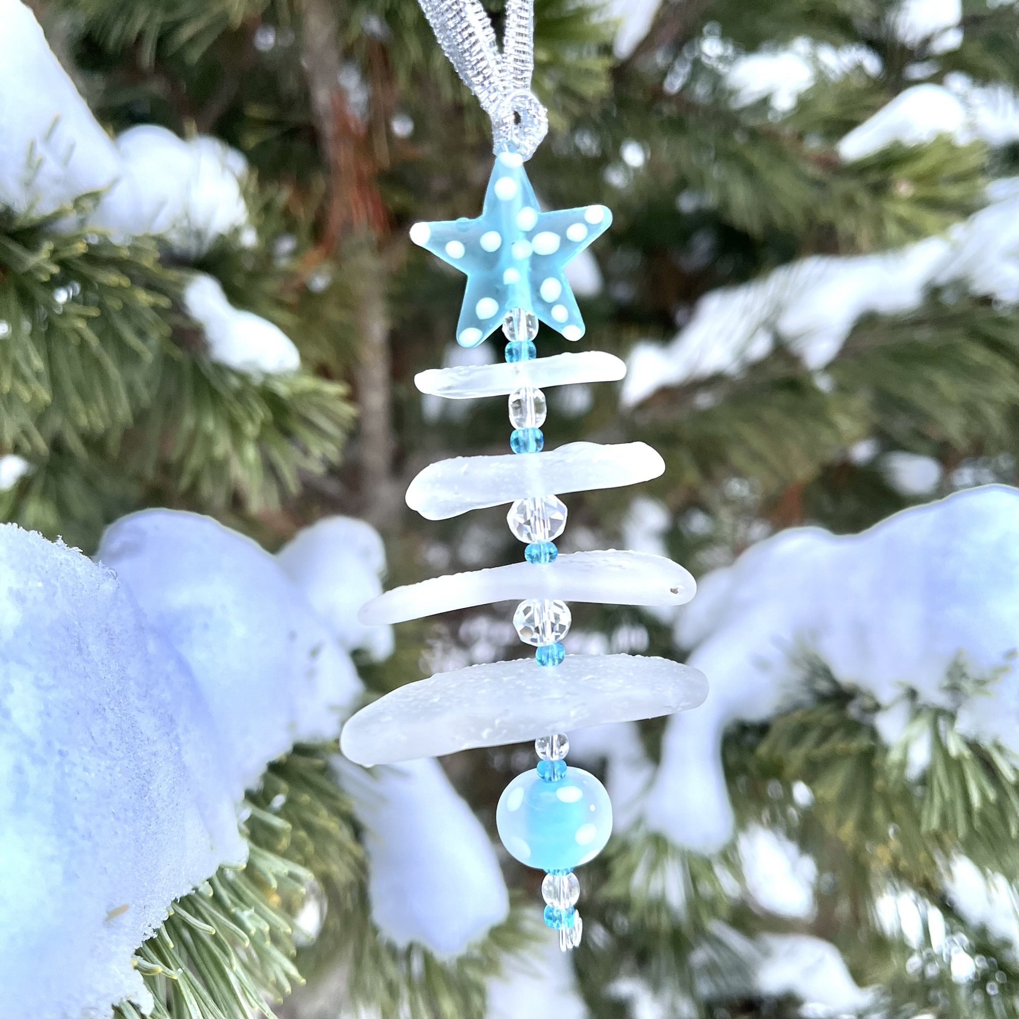 2025 Aroostook River Snow Tree ornament No. 2 hanging outdoors among snowy pine branches.