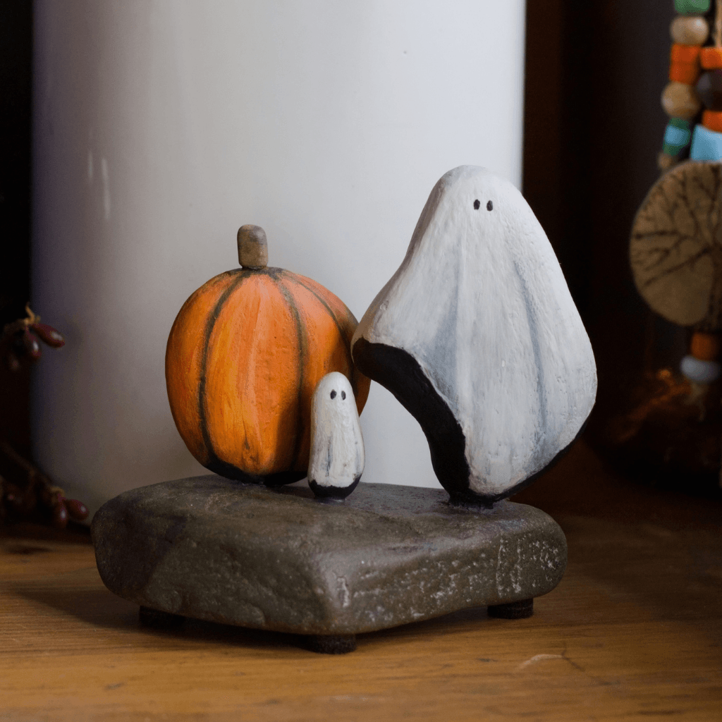 Hand painted stone ghosts and an orange pumpkin displayed on a wooden shelf beside a white vase
