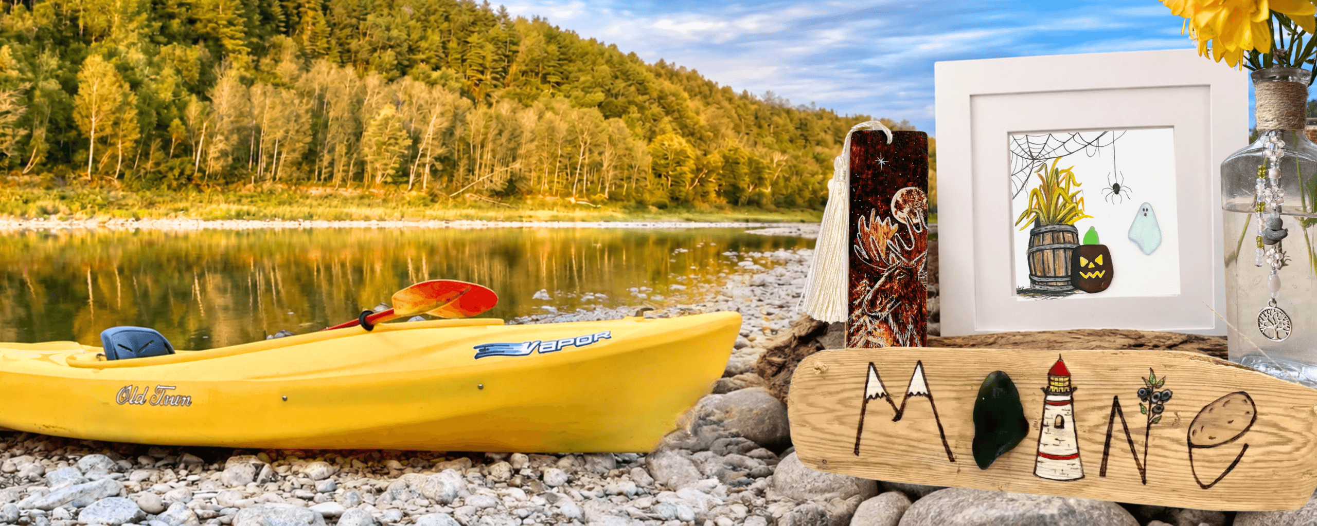 Riverbed Art display beside the Aroostook River in Caribou, Maine, featuring handmade art, a Maine driftwood sign, and yellow kayak.