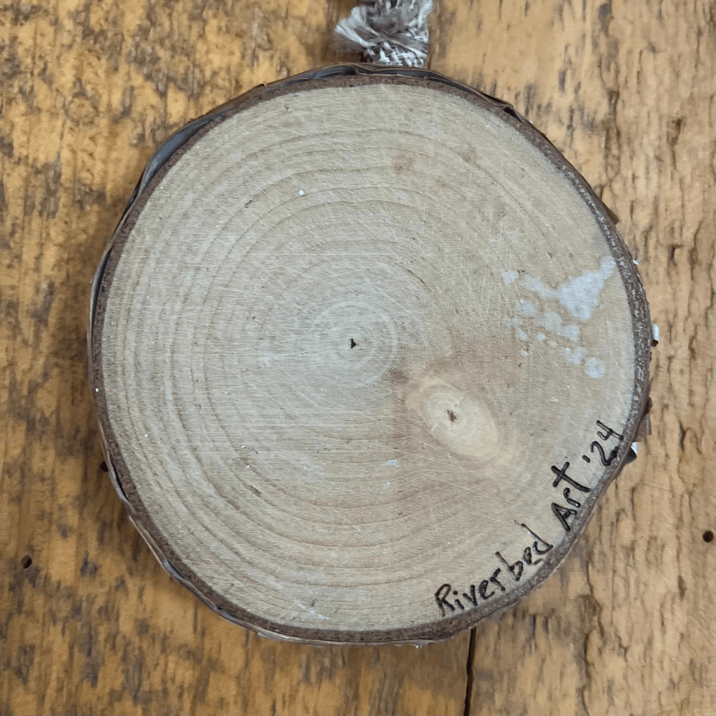 Back of a birch slice ornament showing natural wood rings, bark edge, and the Riverbed Art ’24 signature.
