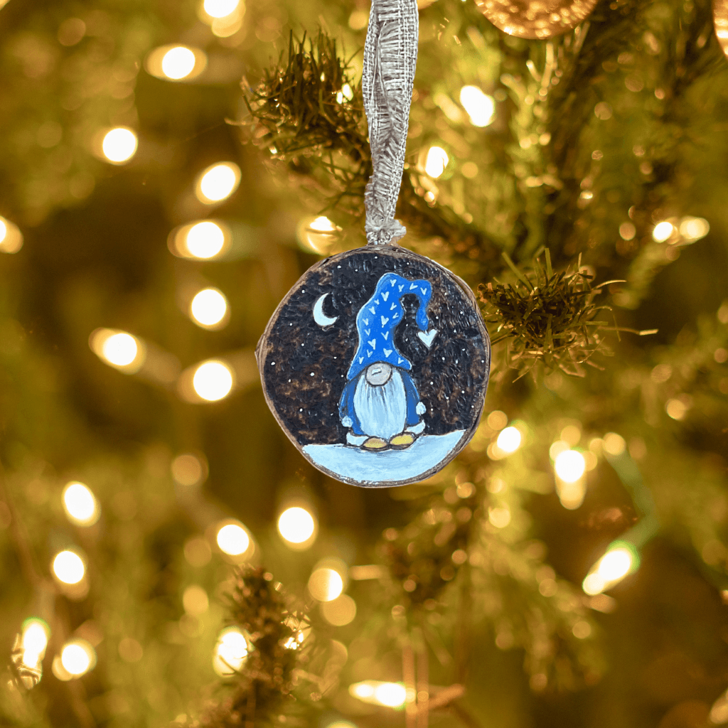 Gnome of Love birch slice ornament hanging from a ribbon among evergreen branches and warm white holiday lights.