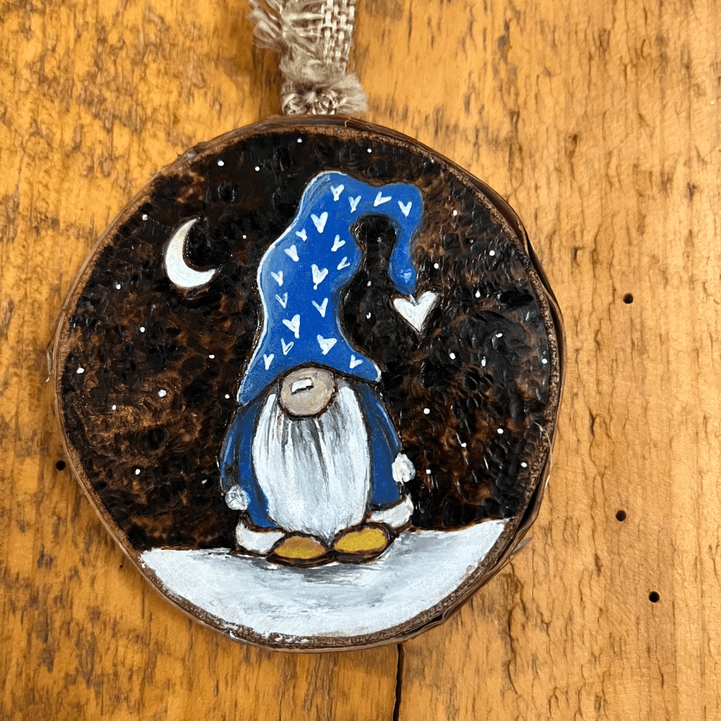 Blue-hatted gnome on a woodburned birch slice with white hearts and a crescent moon, displayed against a wooden wall.