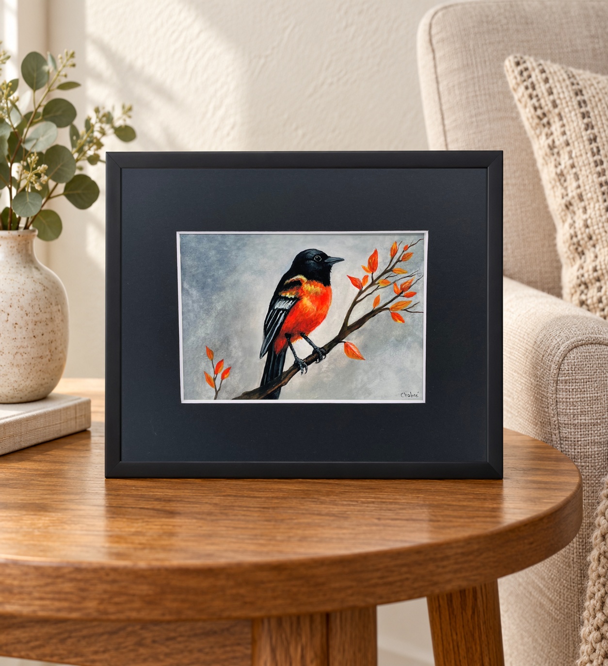 Baltimore Oriole print in a black mat and black frame on an oak side table beside a linen chair and ceramic vase.