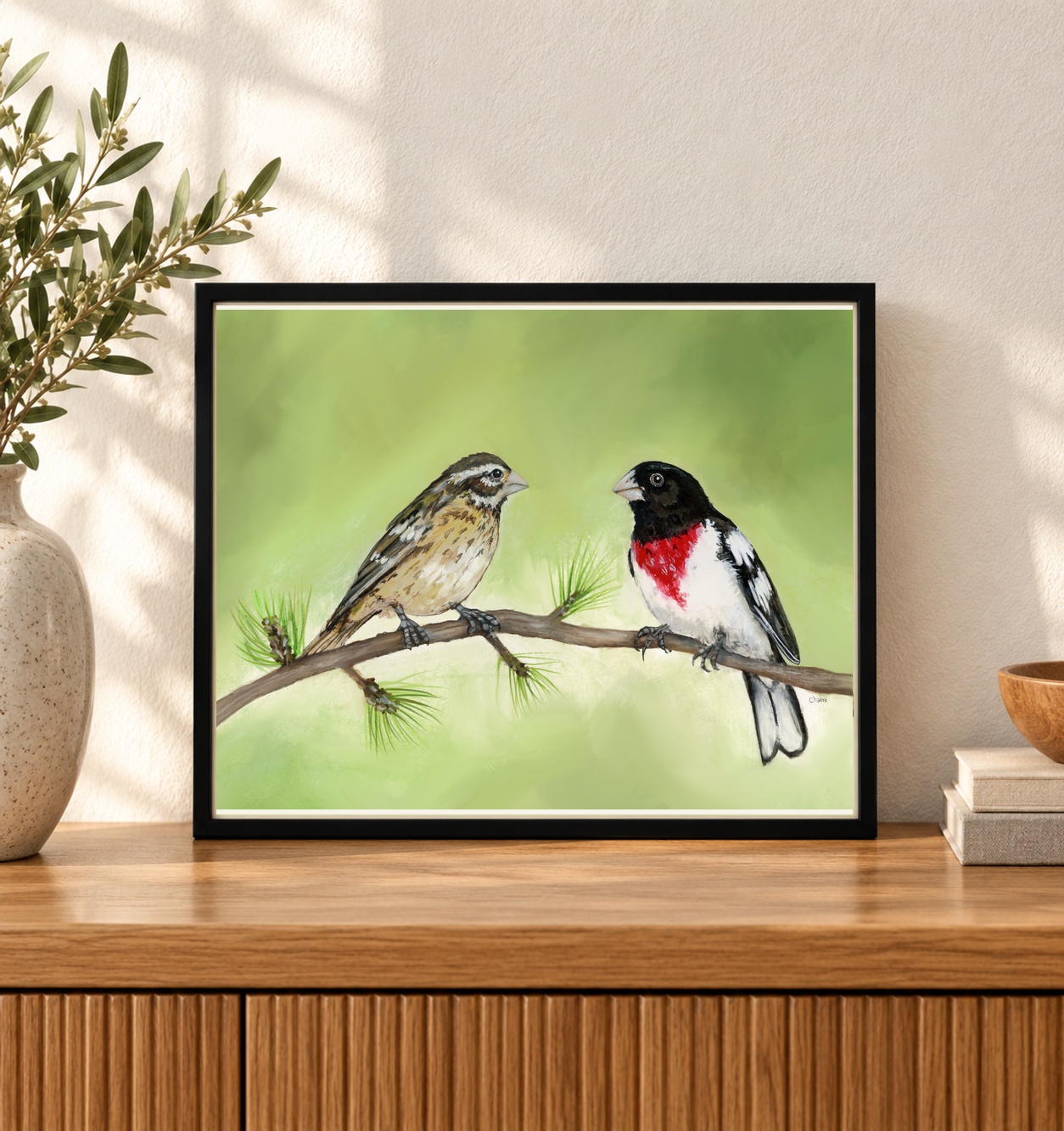 Riverbed Art Grosbeak couple artwork displayed in a black landscape 10 × 8-inch frame on an oak console beside a ceramic vase.