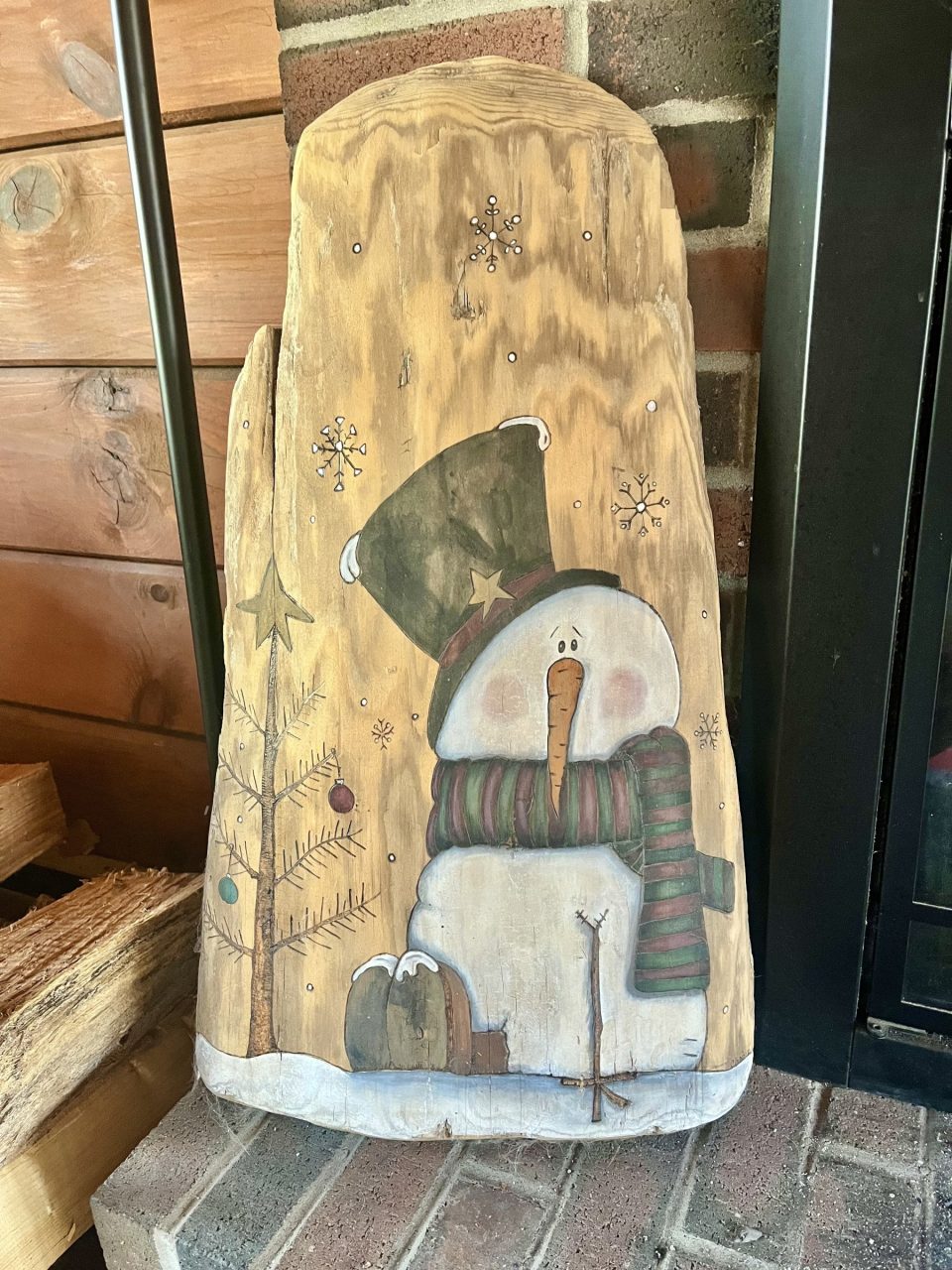 Snowman with a tall hat and striped scarf beside a decorated tree and snowflakes on a wood slab, displayed by a brick hearth